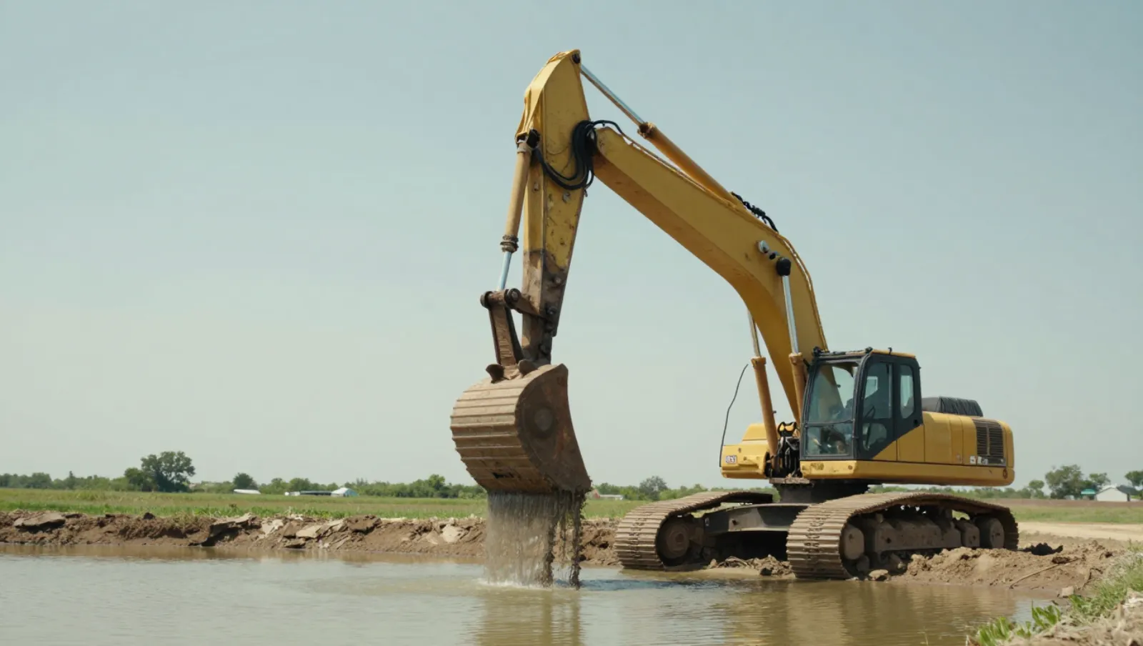 How to Dredge a Pond: Complete Guide 2025 Yellow excavator dumps silt into water on a muddy riverbank while dredging — Daniel Dean Land Clearing, Southeast Texas