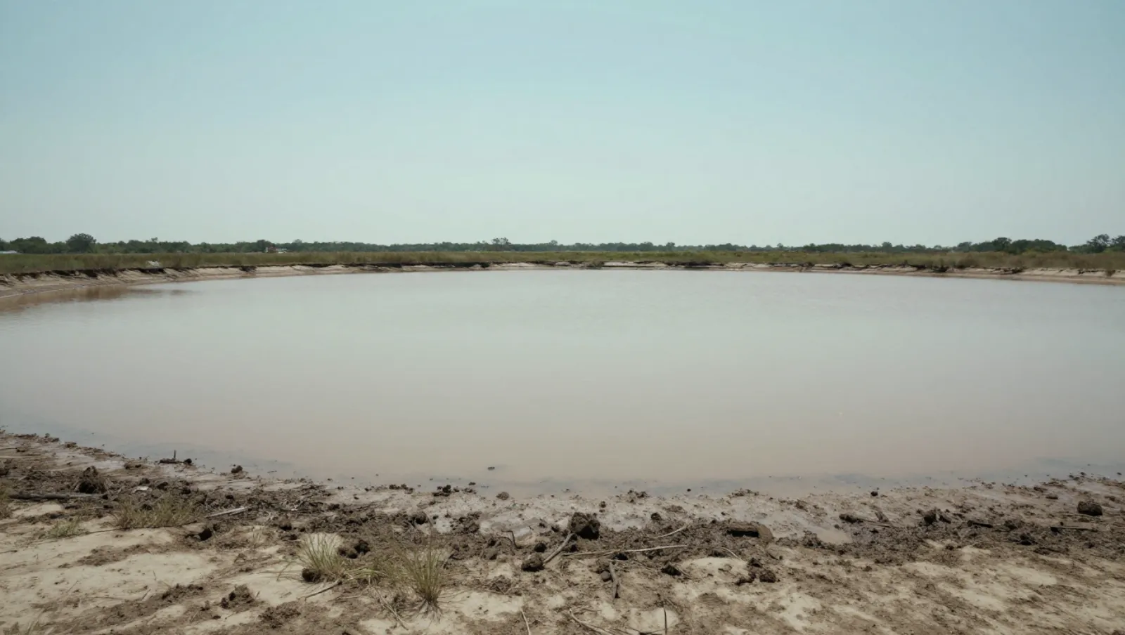 Top 5 Signs You Need Dredging Services for Your Pond or Lake Brown murky water spans a sandy-muddy riverbank with sparse grass; no machinery or work evident — Daniel Dean Land Clearing, Southeast Texas