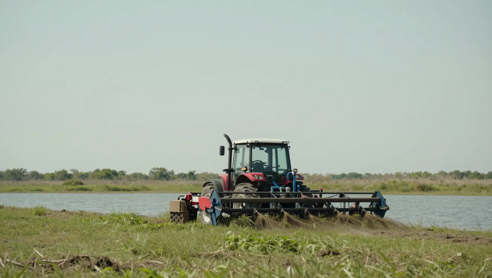 Red tractor on grassy field by water cuts tall vegetation — Daniel Dean Land Clearing, Southeast Texas