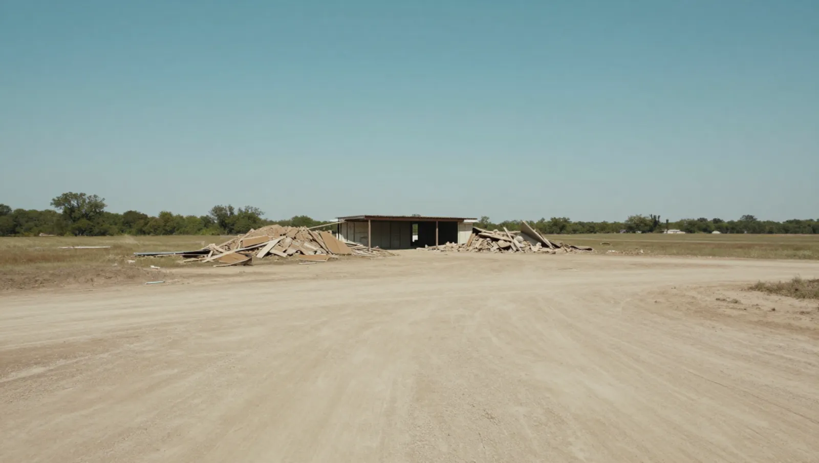Demolition in Pearland, Texas — completed work Wood debris piles show demolition work on a small building across a dusty dirt field; no machinery present — demolition in Pearland, Texas